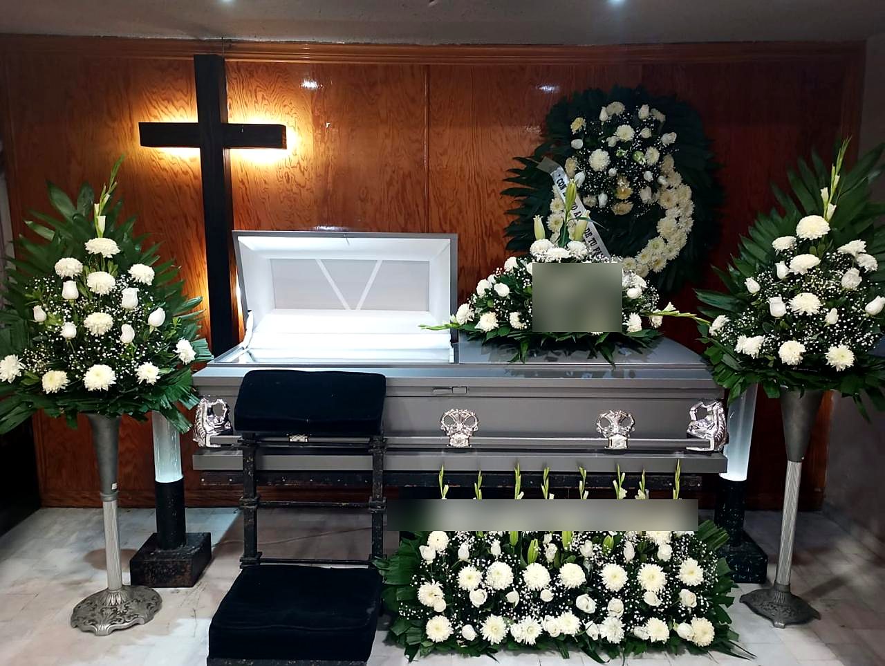 Velación con ataúd plateado, cruz de madera y arreglos de flores blancas